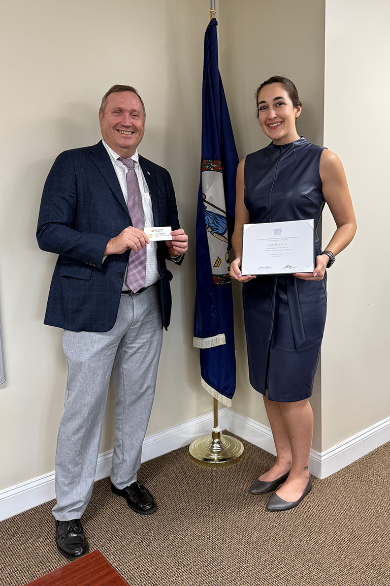 Marina Pugh holds a certificate and stands next to a man in a suit in front of the Virginia flag